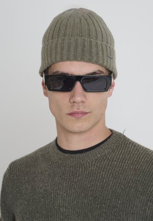 Maslinasto zelena rebrasta pletena kapa i odgovarajući teksturirani pulover. Crne pravokutne sunčane naočale. Jednostavan, strukturiran dizajn s neutralnim bojama.