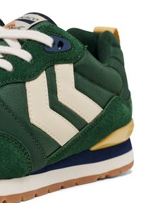 Green sneaker with suede and mesh materials, featuring white chevron accents, blue eyelet, and a yellow accent on the sole.
