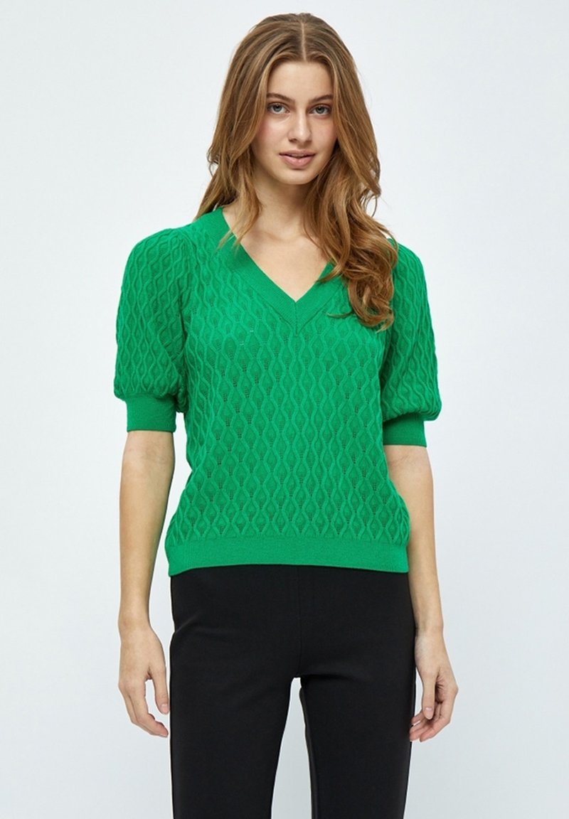 Pull en tricot vert avec un col en V, des manches bouffantes courtes et un motif en texture de diamant, associé à un pantalon noir ajusté.