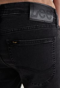Mustat farkut, joissa on Lee-brändin nahkapaikka vyötäröllä, pieni kultainen logo takataskussa ja näkyvät ompelusarjat.