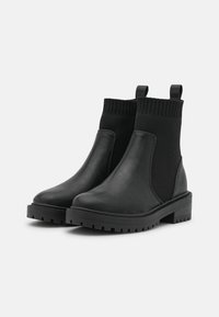 Botines negros hasta el tobillo con exterior de cuero liso, paneles laterales elásticos y un cuello de punto acanalado. Cuentan con una suela de goma gruesa con dibujo de banda de rodadura.