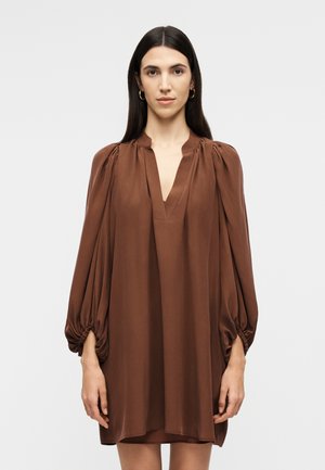 Hosszú, sötét hajú nő, laza, hosszú ujjú barna ruhában, V-nyakú és összegyűrt mandzsettával, egyszínű háttér előtt állva.