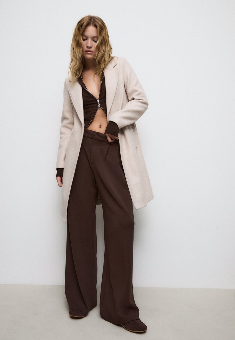 Un manteau en laine beige sur un haut zippé marron et un pantalon large marron, présentant des textures lisses et une coupe décontractée.