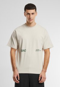 Beige bavlněné tričko s krátkým rukávem, na přední straně s potiskem "SAC" a "Saturday Athletics Club" v zelené barvě. Volný střih.