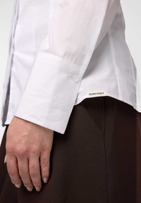 Camicia bianca con un leggero lucido, caratterizzata da un polsino con bottone, indossata sopra pantaloni marroni scuri con un'etichetta visibile che legge "SURI FREY".