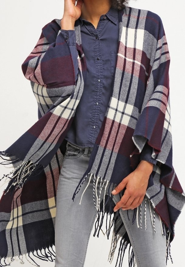 Personne portant une chemise boutonnée bleu marine, un jean gris clair et un grand châle à carreaux bleu marine, bordeaux et blanc avec des franges.