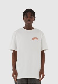 Hvit oversized t-skjorte laget av bomull, med oransje tekstdesign på øvre venstre side. Korte ermer med rund hals.