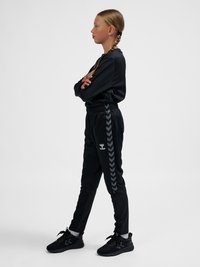 Giovane ragazza con capelli intrecciati in piedi con le braccia incrociate, indossa abbigliamento sportivo nero con pantaloni a righe a spina di pesce e scarpe da ginnastica nere su uno sfondo semplice.