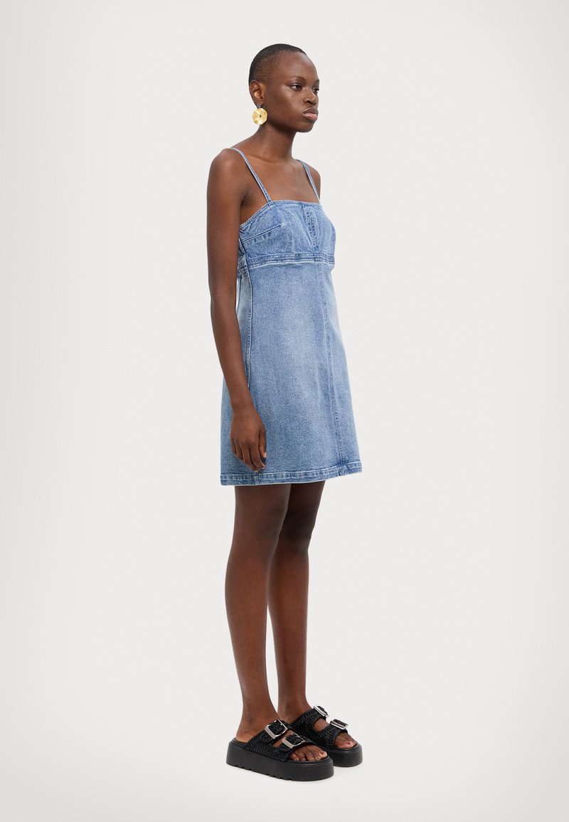 Denimkjole med tynne stropper, en tettsittende liv og utsvingt skjørt. Har blå farge, synlig søm og lengde til midten av låret. Svarte platåsandaler.