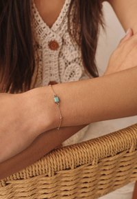 Gold chain bracelet featuring a turquoise square pendant, smooth surface, delicate links, worn on a wrist against a woven background.