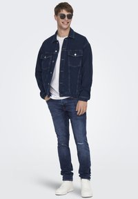 Jachetă din denim de culoare bleu închis, cu buzunare frontale și închidere cu nasturi, purtată peste un tricou alb. Blugi bleu deschis și adidași albi.
