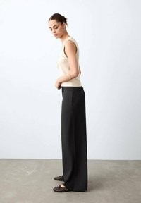 Femeie stând lateral, purtând un top bej fără mâneci, pantaloni negri cu talie înaltă și croială largă, și pantofi platați maro închis.