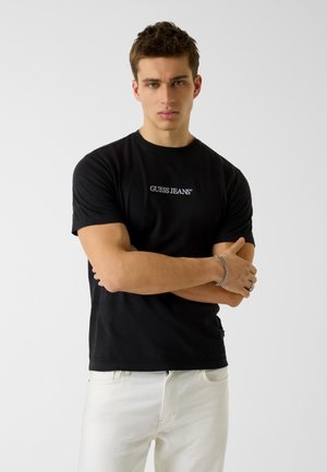 Giovane uomo con capelli corti che indossa una t-shirt nera con la scritta "GUESS JEANS" e pantaloni bianchi, in piedi con le braccia incrociate davanti a uno sfondo semplice.