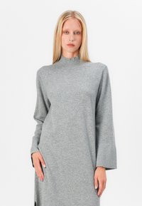 Siva dolgo-sleeve obleka iz mehkega materiala z visokim ovratnikom, z širokimi rokavi in stranskimi razporki za dodaten učinek.