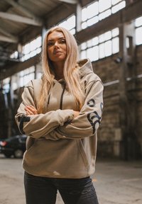 Sweat à capuche beige avec une poche avant et un logo noir sur la manche, associé à un jean noir déchiré. Cadre industriel intérieur.