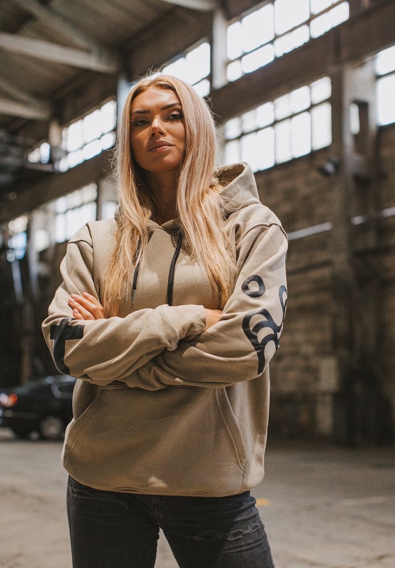 Sweat à capuche beige avec une poche avant et un logo noir sur la manche, associé à un jean noir déchiré. Cadre industriel intérieur.