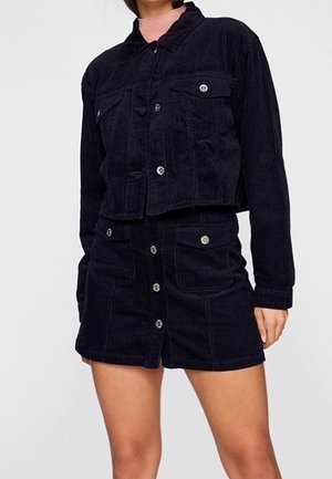 Femme portant une veste en velours côtelé noir à boutons et une mini-jupe assortie à boutons sur le devant avec poches avant, debout devant un fond uni.