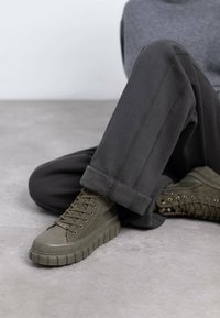 Zapatillas altas verdes de ante y goma, con una suela gruesa y texturizada y ojetes plateados, combinadas con pantalones gris oscuro.