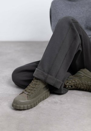 Zapatillas altas verdes de ante y goma, con una suela gruesa y texturizada y ojetes plateados, combinadas con pantalones gris oscuro.