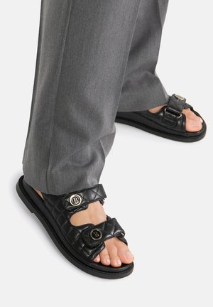 Czarne pikowane sandały z dwoma regulowanymi paskami oraz okrągłymi metalicznymi ozdobami, zestawione z szarymi, dopasowanymi spodniami.