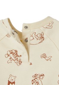 Cremfarbener Oberteil aus weichem Stoff mit braunen Cartoon-Tierdruck. Verfügt über einen gerippten Halsausschnitt und zwei Druckknöpfe am Kragen.