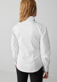 Camicia bianca a maniche lunghe con un design aderente, colletto alto e spalle a sbuffo arricciate. Tessuto liscio e leggermente lucido con polsini a bottoni.