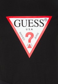 Černé tričko s velkým červeným a bílým trojúhelníkovým logem s textem "GUESS U.S.A." a "WASHED JEANS" a otazníkem uprostřed.