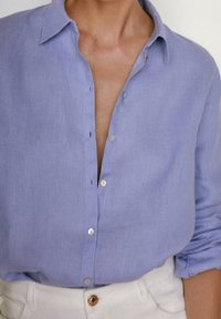 Camicia in lino viola chiaro con colletto classico, caratterizzata da chiusura con bottoni e maniche arrotolate, abbinata a una cintura di colore chiaro.