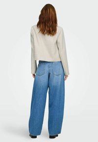 Donna con capelli castani di media lunghezza, indossa una giacca grigio chiaro e jeans blu a gamba larga, in piedi di spalle su uno sfondo neutro.