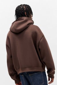 Felpa marrone oversize con tessuto strutturato, cappuccio ampio e polsini a costine. Abbinata a denim scuro, vista da dietro.