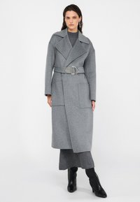 Šedý vlnený kabát s výstrihom, pásom na stiahnutie, dvoma prednými vreckami a dĺžkou po kolená. Nosený cez šedý outfit a čižmy.