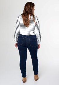 Jean skinny bleu foncé avec une taille haute, associés à une chemise blanche à rayures à manches longues. Le tissu semble lisse ; la chemise a une légère bouffante dans les manches.