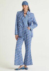 Completo blu a motivo con pantaloni svasati e blazer. La maglietta presenta un disegno geometrico. Accessoriato con una sciarpa coordinata e scarpe basse.