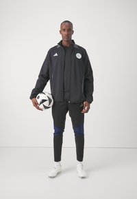Chaqueta deportiva negra con emblema verde y blanco, combinada con pantalones negros que tienen acentos azules, sosteniendo un balón de fútbol blanco y negro, con zapatillas blancas.