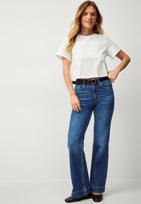 Maglietta bianca corta abbinata a jeans flare blu in denim a vita alta e una cintura nera. I jeans presentano un classico design a cinque tasche e orlo dritto.