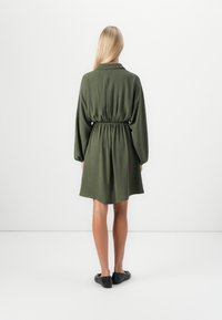 Robe chemise verte ceinturée, à manches longues, coupe décontractée et tissu texturé. Fini au milieu des cuisses, associée à des chaussures noires à enfiler.