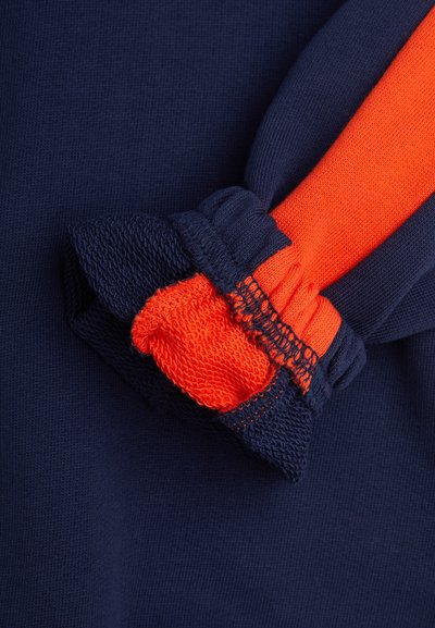 Temno modra obleka z oranžnim poudarkom, z nabranimi manšetami, ki prikazujejo teksturirano pletenino in gladek zaključek.