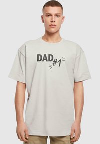 Lichtgrijs t-shirt met korte mouwen en een ontspannen pasvorm, voorzien van zwarte opdruk "DAD #1" en grafische accenten op de voorkant.