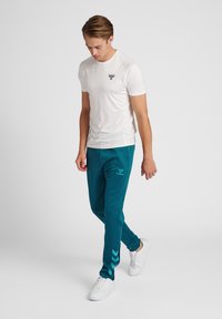 Witte sportt-shirt met een klein embleem, gecombineerd met teal joggers met een logo en chevronpatroon aan de zijkanten. Witte sneakers.