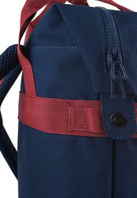 Mochila de tela marinera con correas rojas, que cuenta con un cierre de cremallera, superficie texturizada y un acento de botón negro redondo.