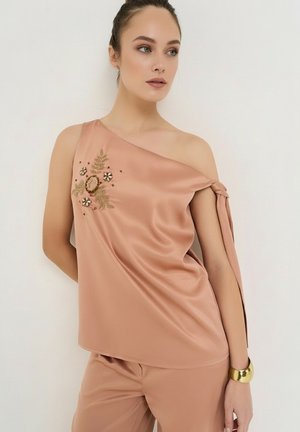 Ung kvinde iført en ferskenfarvet satin top med ét skulderstykke med blomsterbroderi og matchende bukser, accessoriseret med et guldbånd.