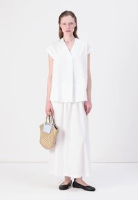 Haut en lin blanc à manches courtes et en V, associé à une longue jupe blanche. Porte un petit sac en osier. Chaussures plates noires.