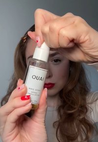 Heldere glazen sprayfles gelabeld met "OUAI WAVE SPRAY," voorzien van een witte pompdeksel. De vloeistof erin is geelachtig. Hand die de fles stevig vasthoudt.