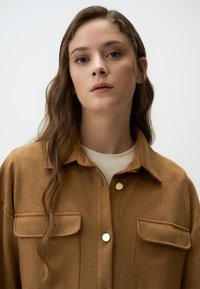 Veste chemise en suède marron avec deux poches poitrine, boutons dorés et coupe décontractée. Modèle portant une chemise de couleur crème en dessous.