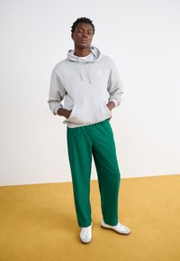 Hallitusseene halli kapuutsiga sviit logoga, millele on lisatud roheline mustriga treeningpüksid. Mudel kannab valgeid spordijalatsid kollasel taustal.