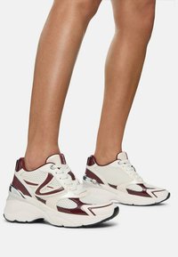 Baskets de sport blanches et bordeaux avec une tige en maille, un col rembourré et des accents réflecteurs. Semelle texturée et lignes de design visibles.