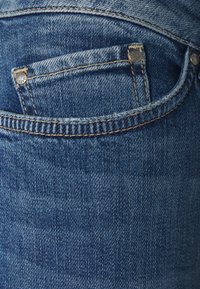 Vaqueros de mezclilla azul con un bolsillo trasero que presenta remaches plateados y costuras visibles, mostrados en primer plano con una apariencia de tela texturizada.