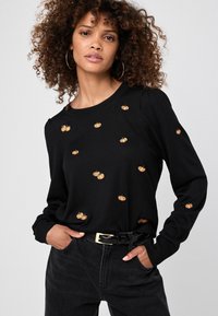 Maglione nero con maniche lunghe, caratterizzato da ricami di zucche oro e arancio. Polsi e orlo a coste; indossato con jeans scuri e una cintura nera.