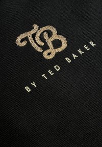Guld broderet "TB" logo over hvid "BY TED BAKER" tekst på sort stof baggrund.
