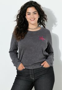 Šedá dlouhá rukávová košile s texturovaným designem hvězdy z flitrů v růžové a modré barvě na přední straně, spárovaná s tmavými džíny.